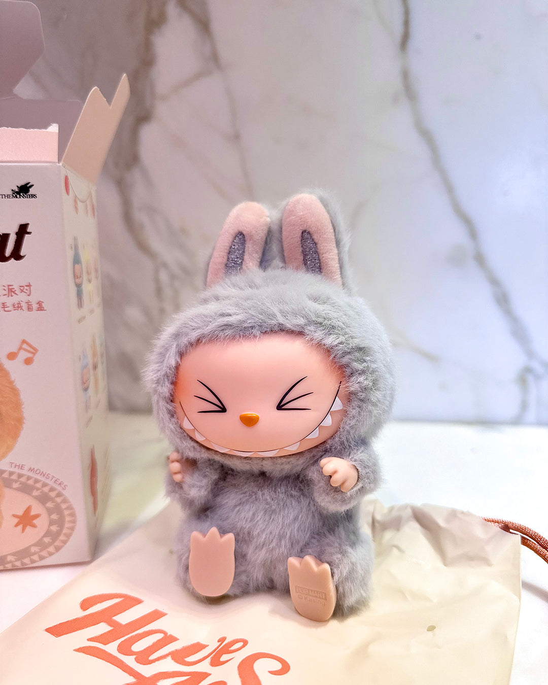 Small plush toy with a gray hood and pink face on a white surface with a blurred background