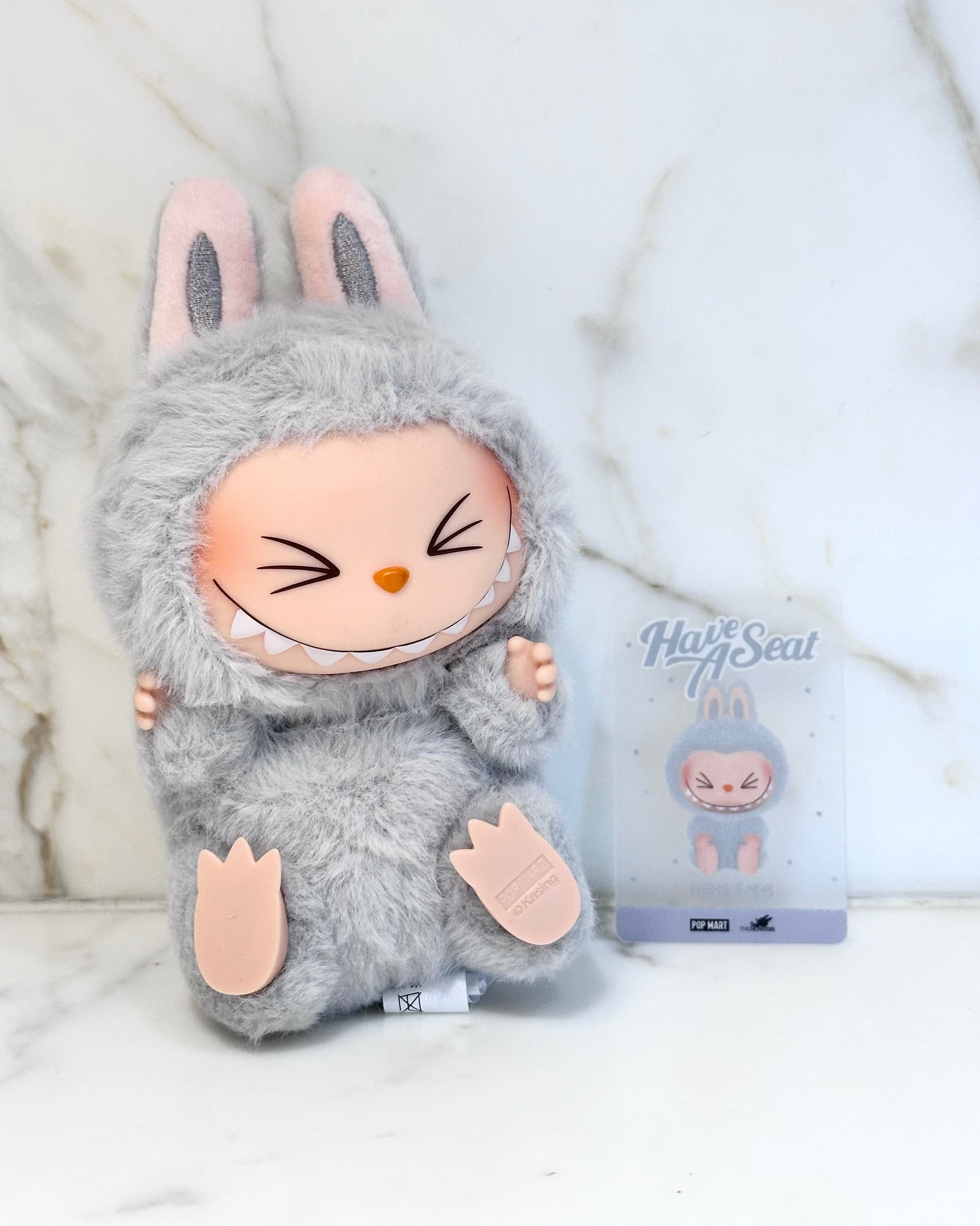 Gray plush toy with bunny ears and a package labeled 'Hug A Seal' on a white background