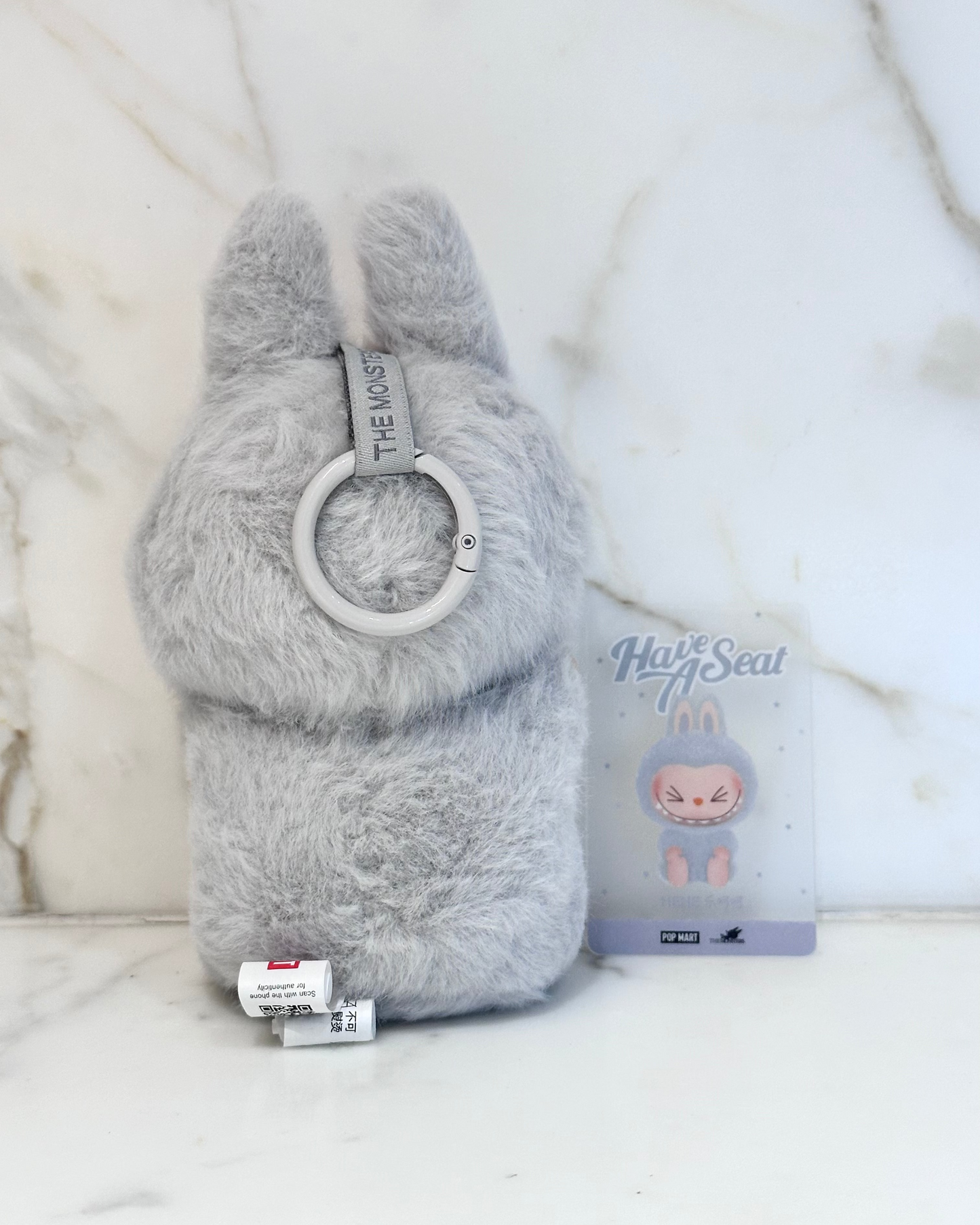 Gray plush toy with a ring and packaging on a white background