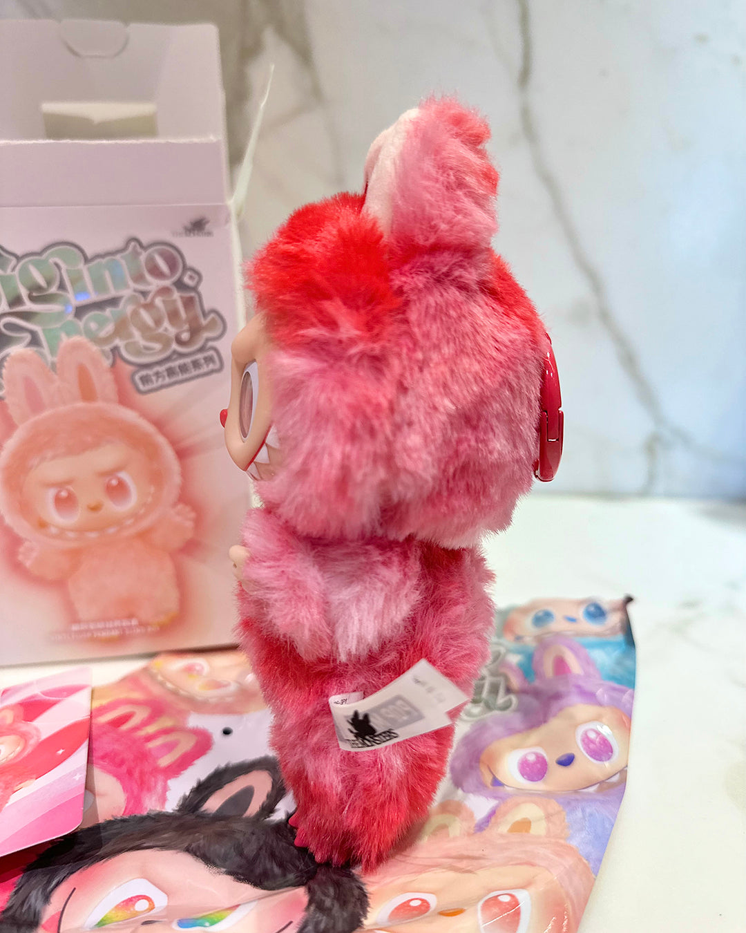 Pink plush toy with a tag on a white surface with cartoon character posters.
