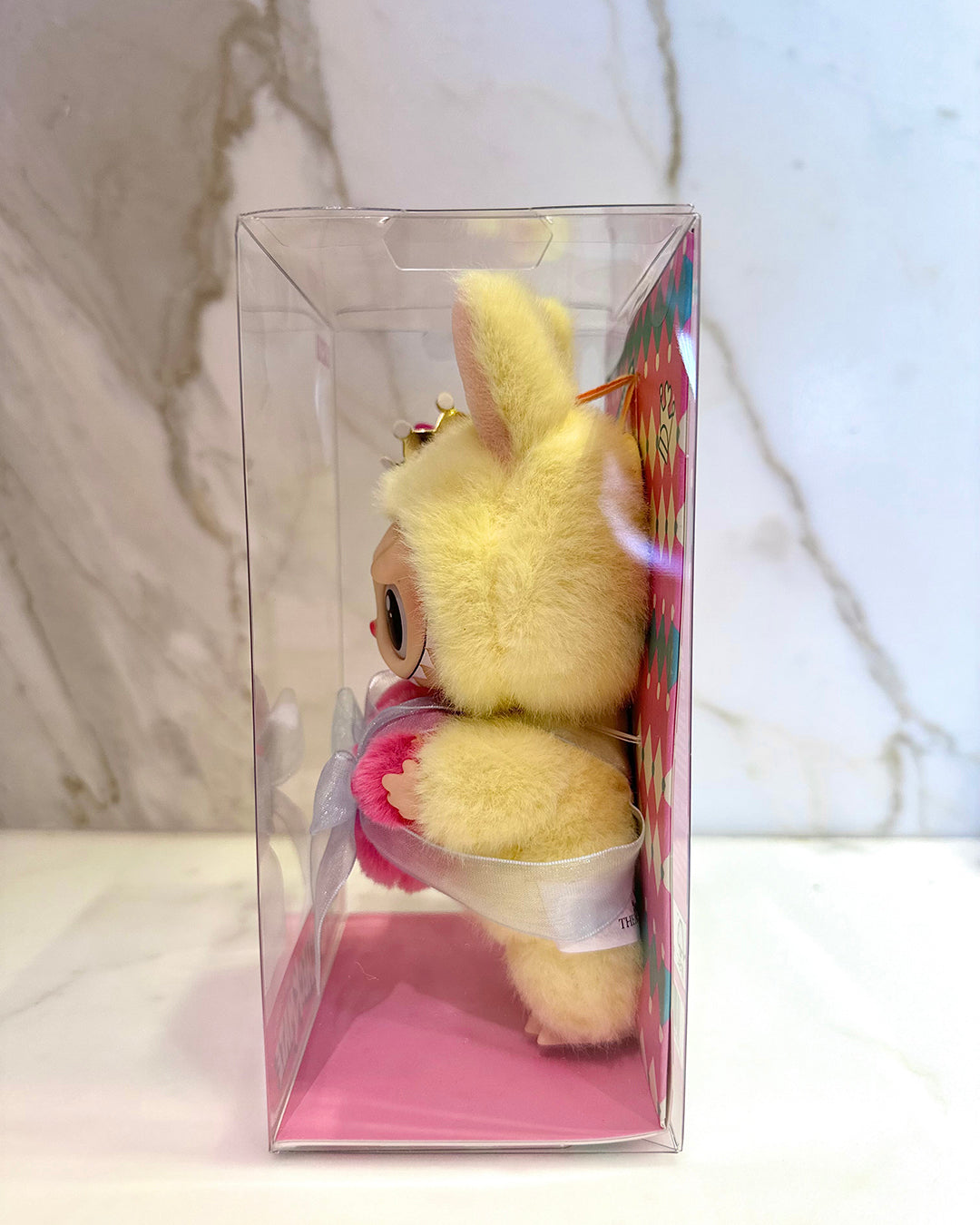 Plush toy in a clear packaging box with a pink interior on a marble surface.