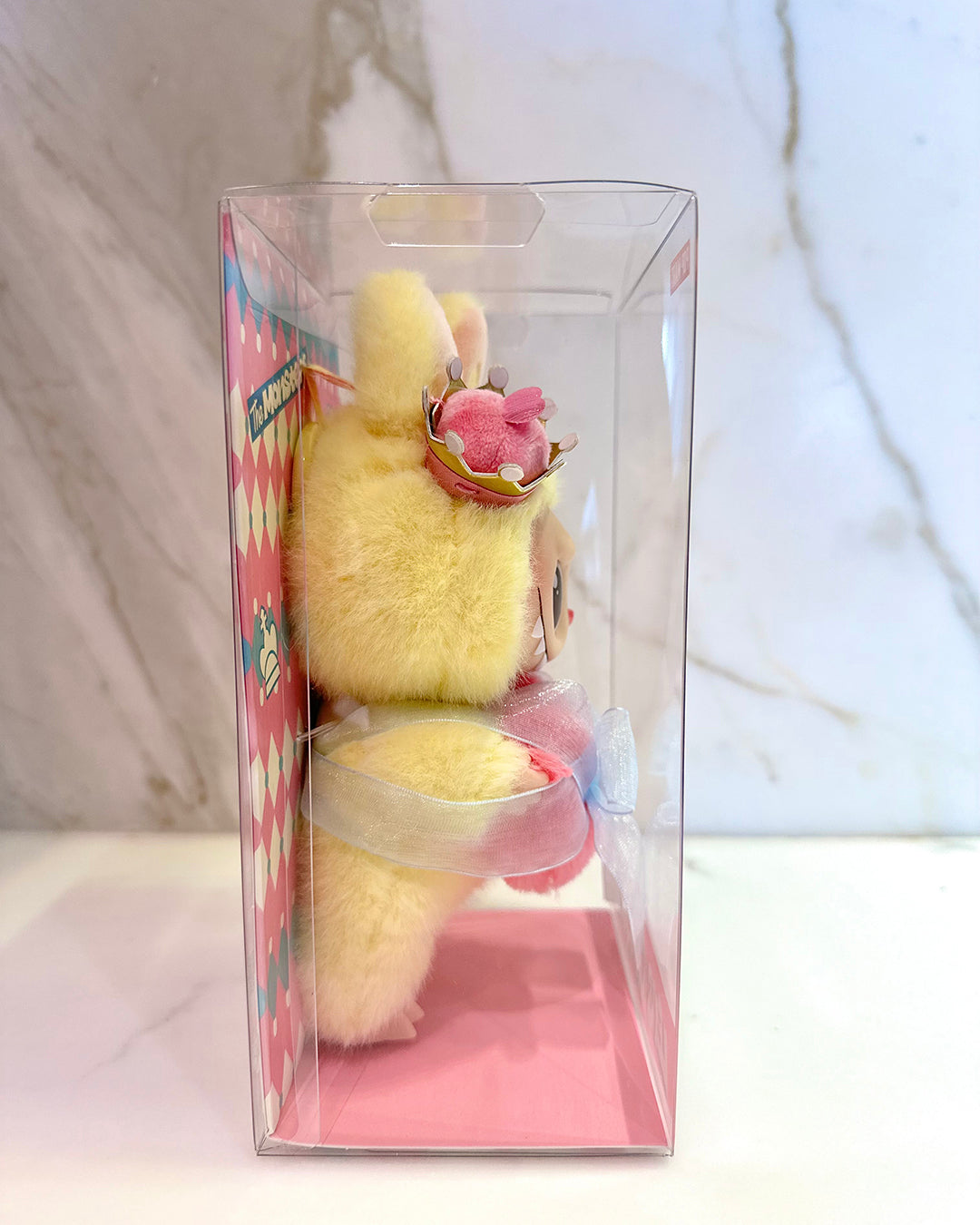 Yellow plush toy with a pink bow in a clear packaging box on a white background