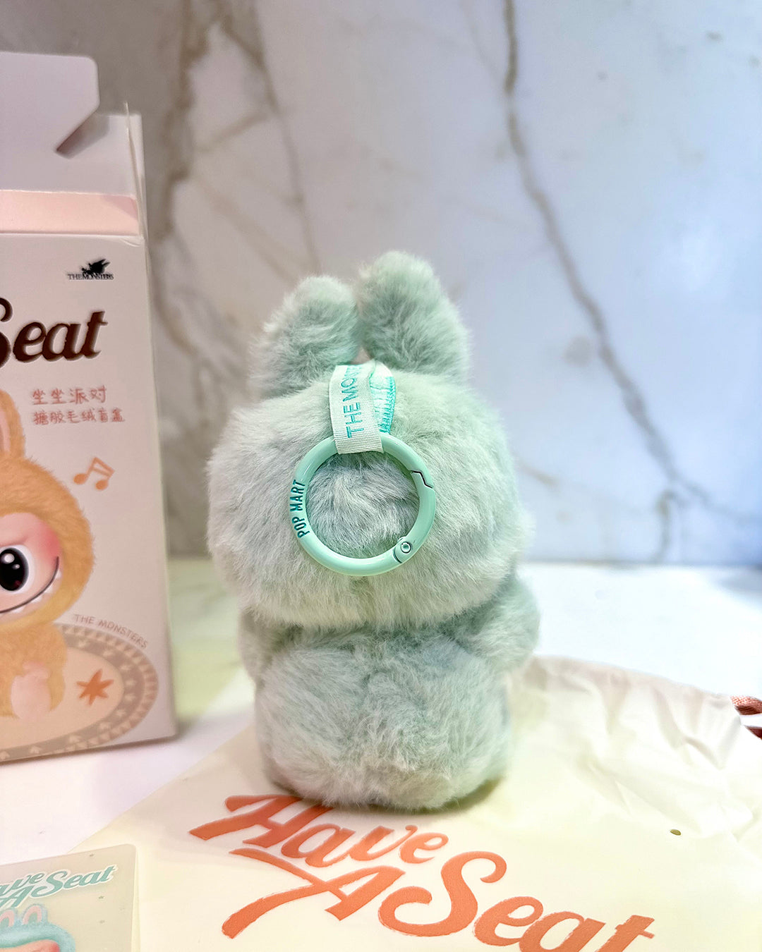Plush toy with a green ring on top of a box labeled 'Have A Seat' with a blurred background.