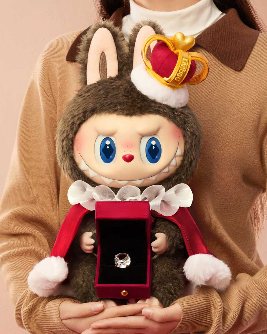Toy figure with a crown and blue eyes holding a diamond ring box, held by a person in a brown coat.