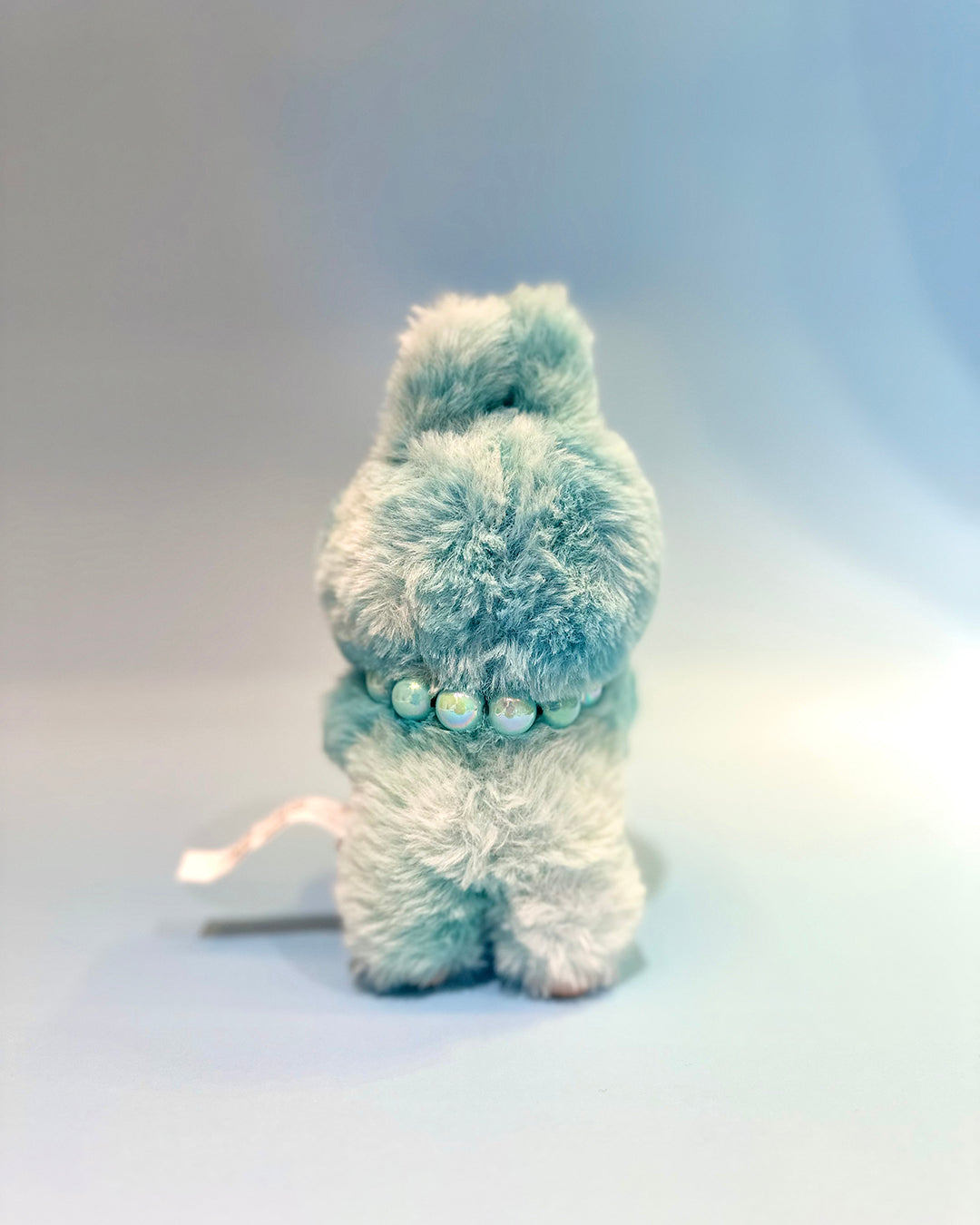 Small plush toy with a fluffy texture and pearl-like beads on a light gray background