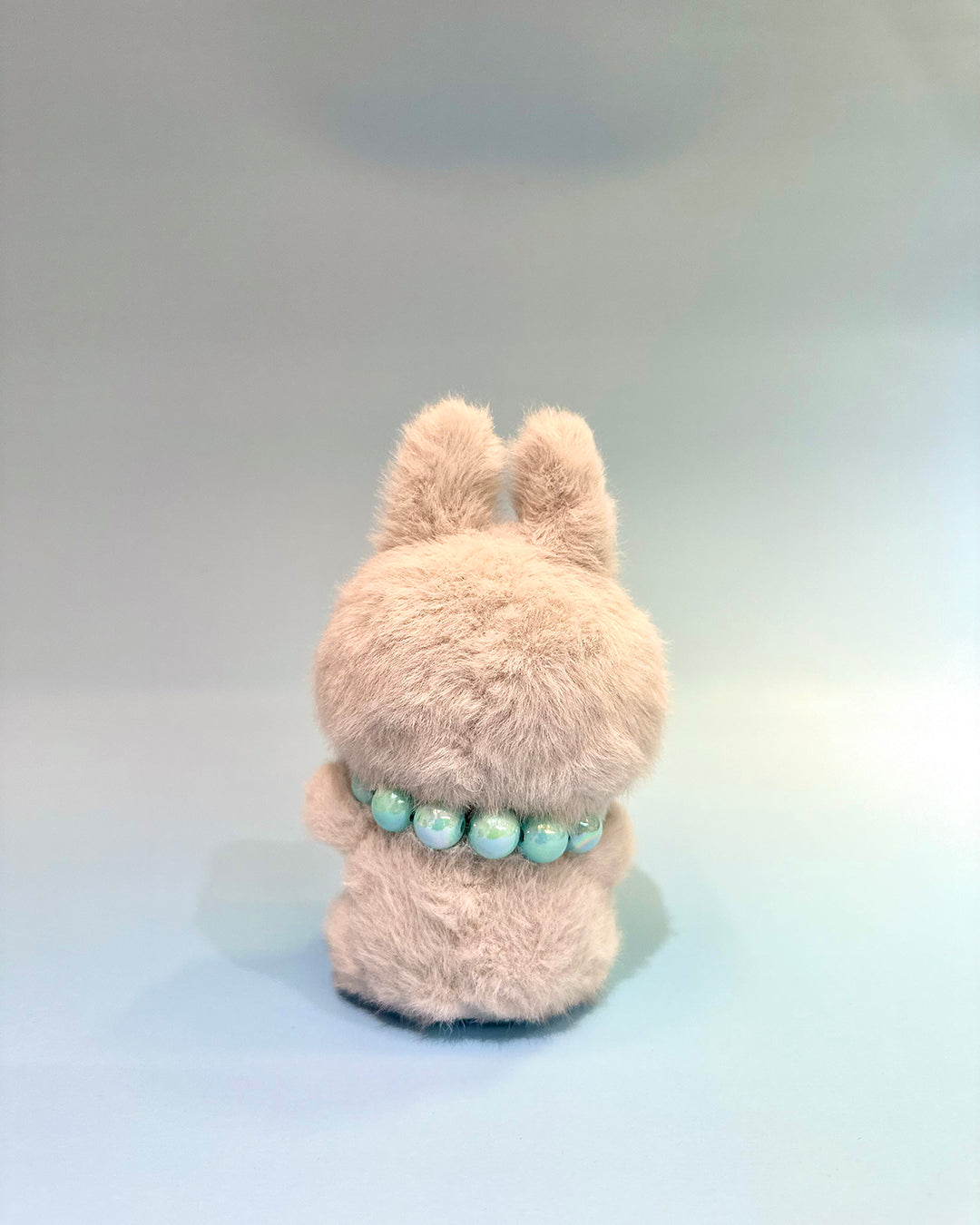 Fluffy beige plush toy with a green beaded necklace on a light gray background