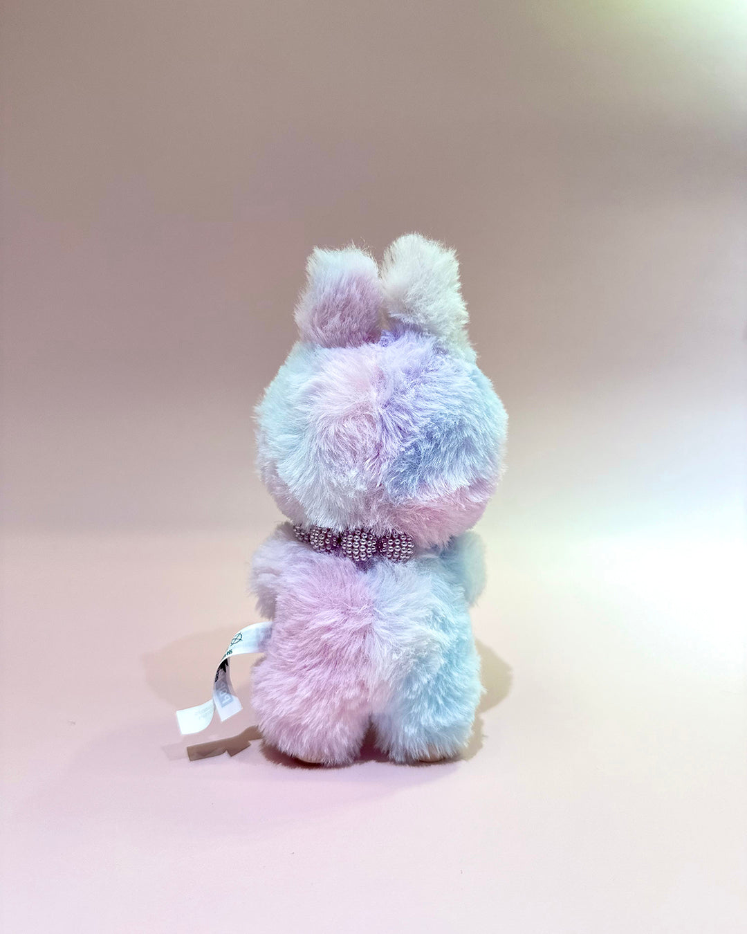 Pastel-colored plush toy with a decorative ribbon on a light pink background