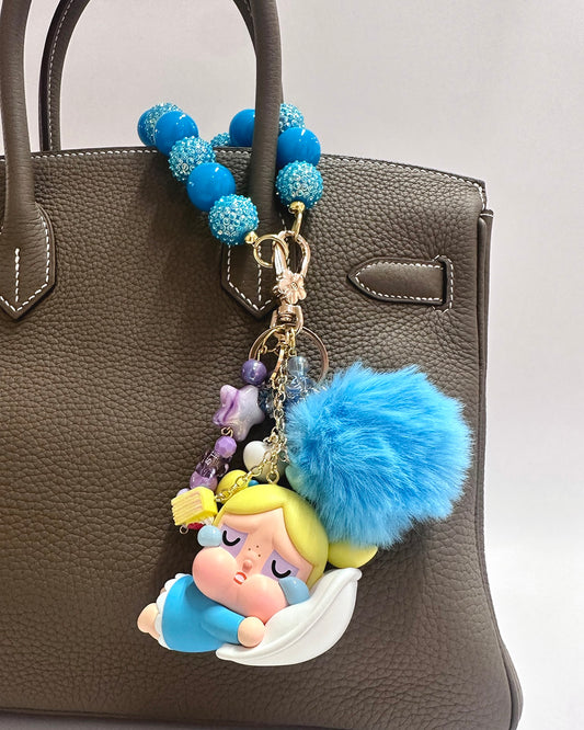 Decorative keychain with blue fluffy ball and cartoon character on a brown handbag.