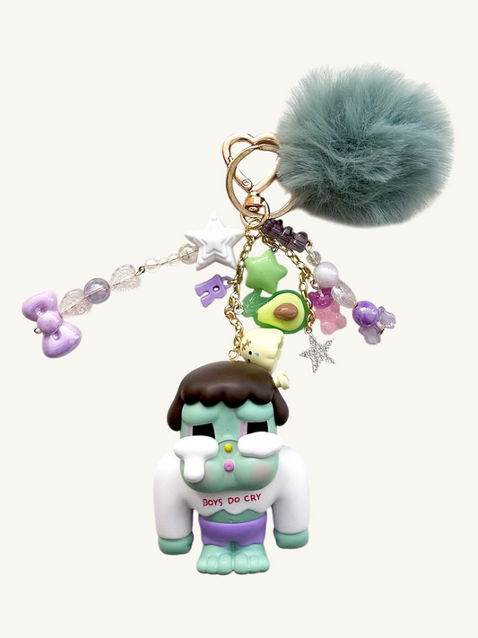 Keychain with character figure, avocado charm, and fur ball on a white background