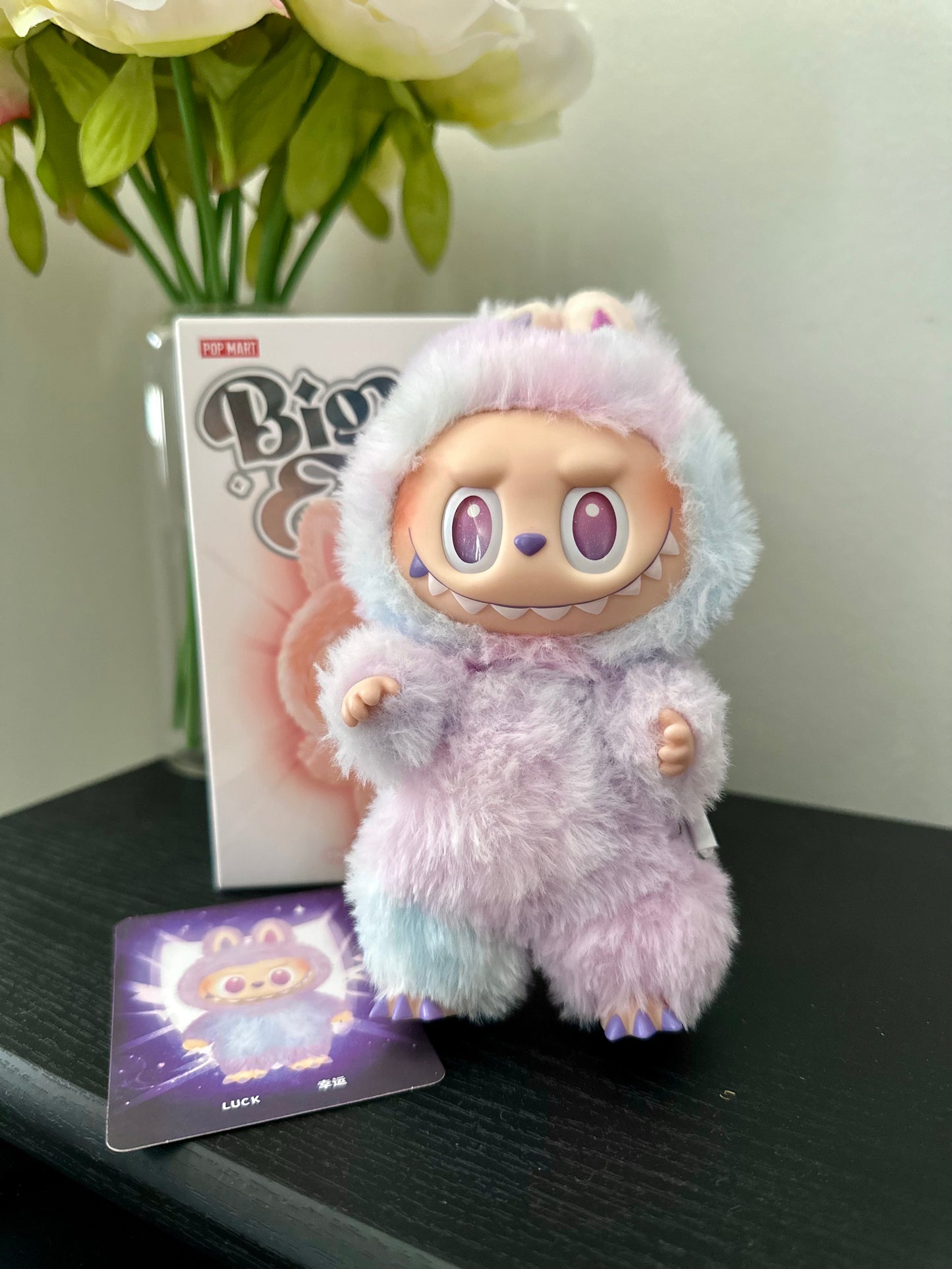 Plush toy with pastel fur and large eyes on a dark surface with a card and flowers in the background.
