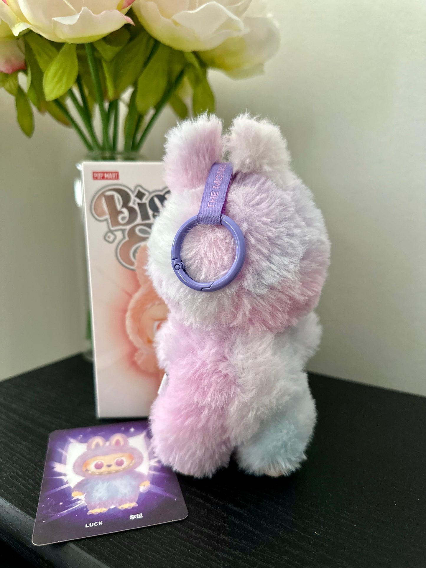 Fluffy pink and white keychain with a purple ring on a dark surface with flowers and a box in the background.