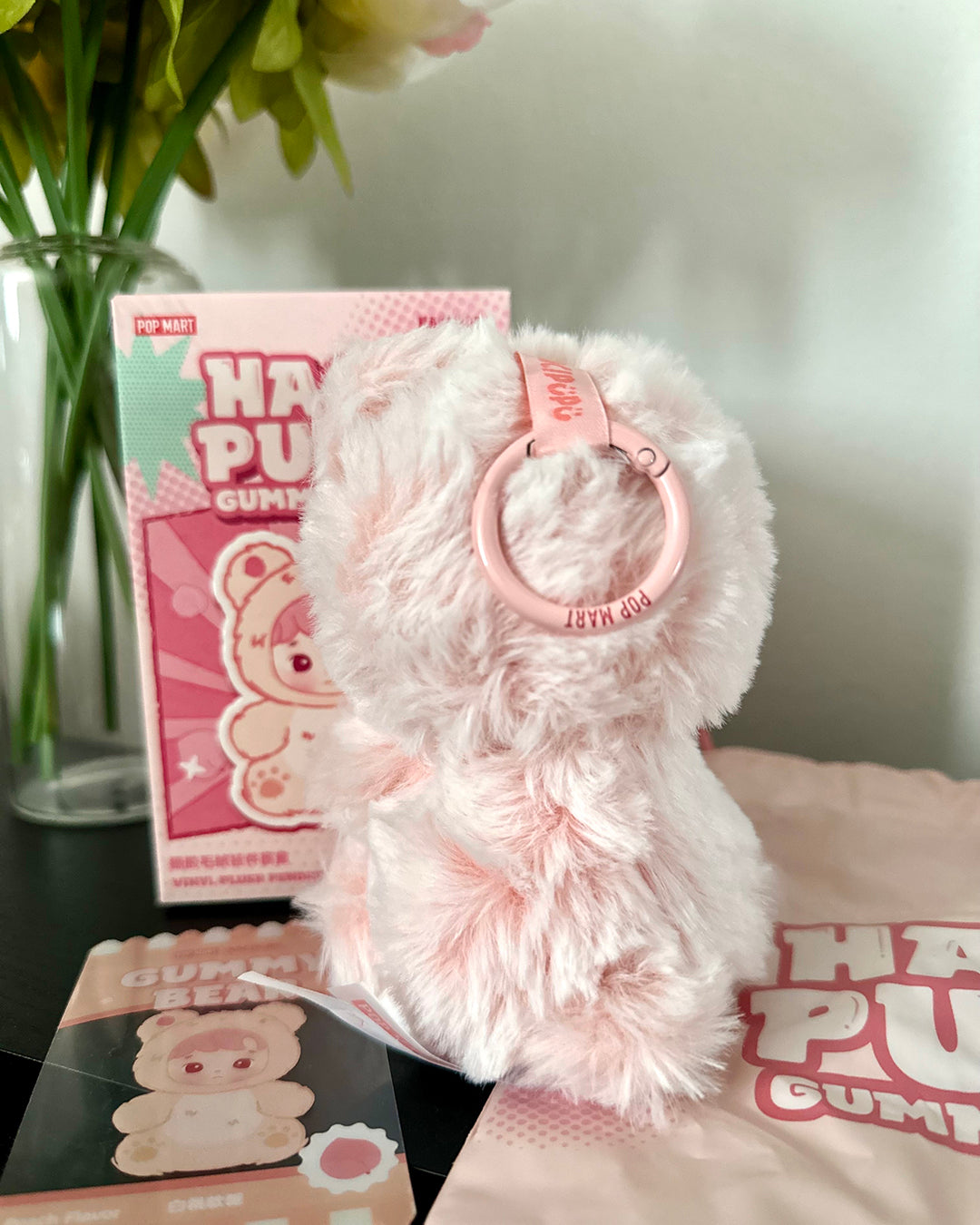 Fluffy pink keychain with 'Happy Plush' branding on a surface with packaging and flowers in the background.