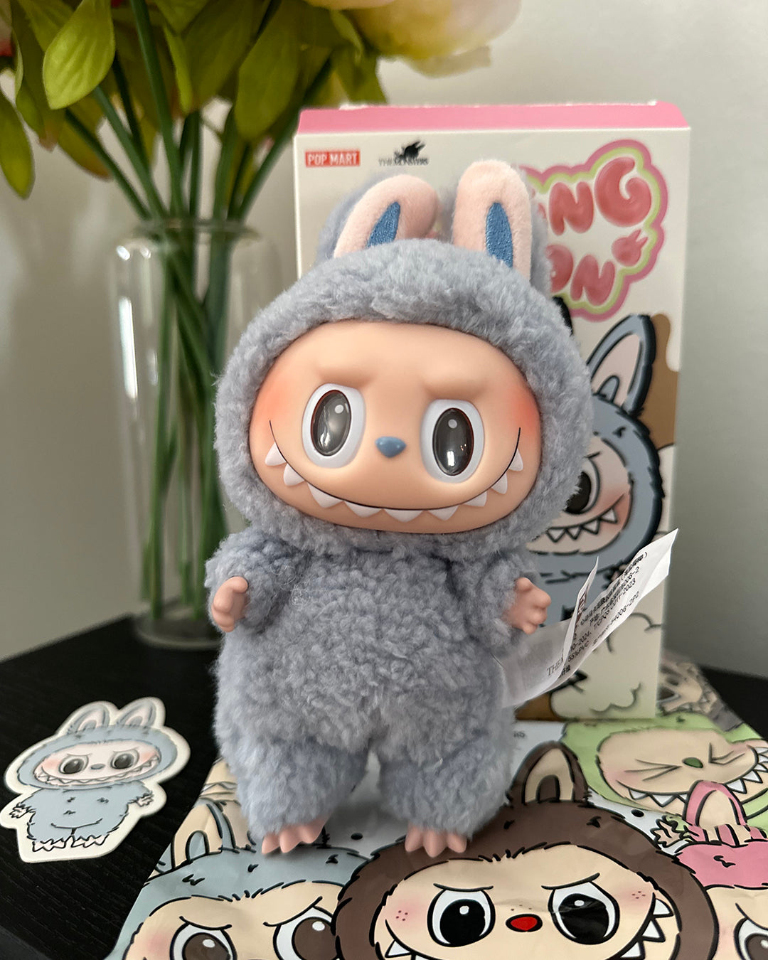 Plush toy in a gray costume with bunny ears on a surface with packaging and stickers in the background.