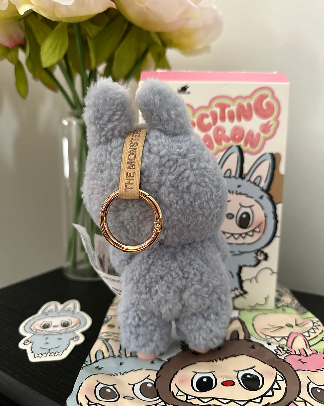 Fluffy gray keychain with a gold ring on a surface with a box and flowers in the background.