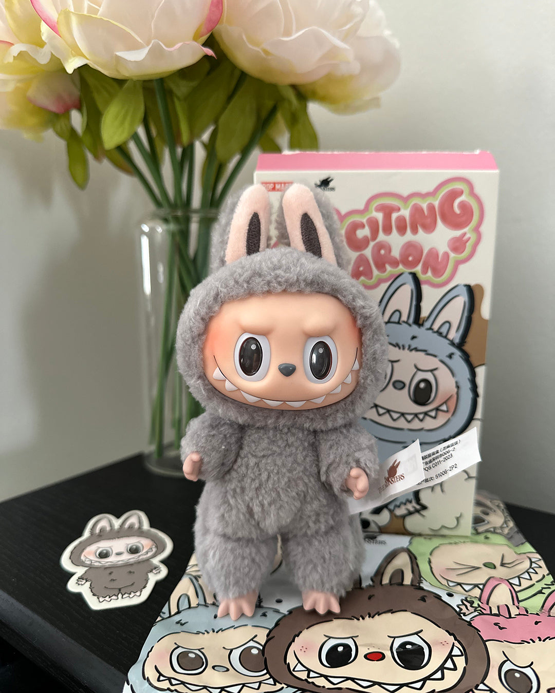 Plush toy in a gray costume with a box labeled 'Citing Aron' and flowers in the background.