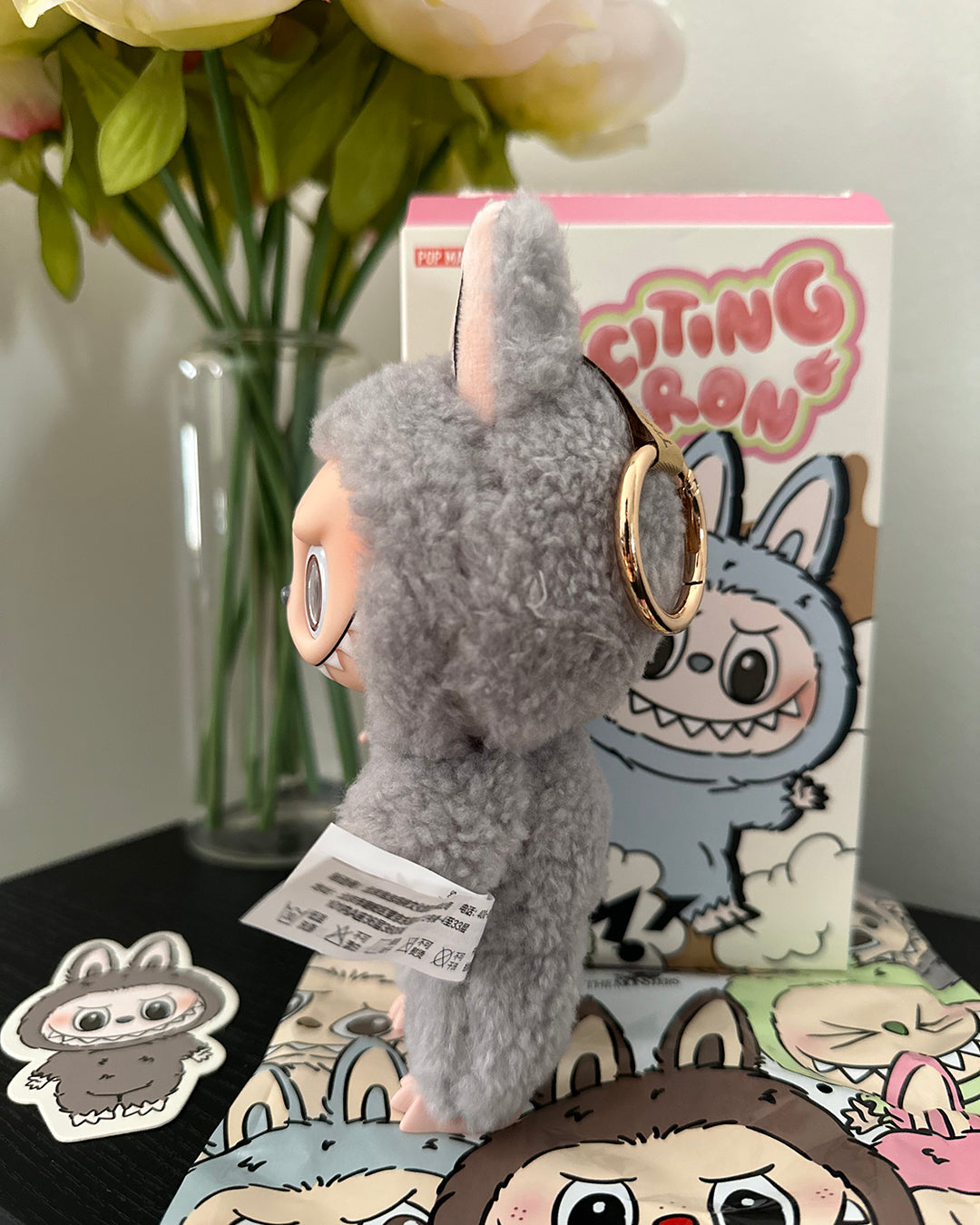 Gray plush toy with a keychain, standing next to a box featuring cartoon characters, with flowers in the background.