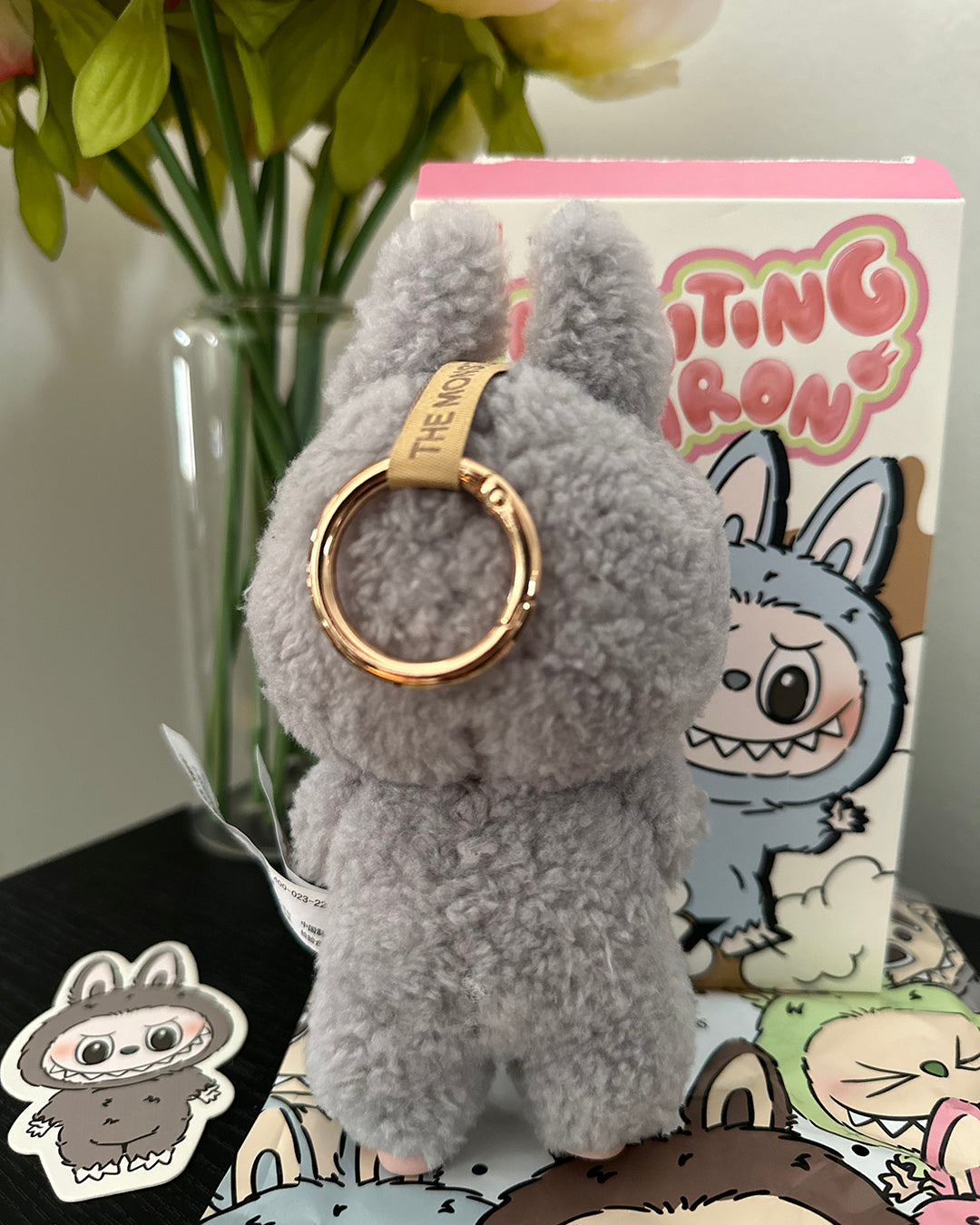 Gray plush keychain with a gold ring on a surface with a cartoon character box in the background.