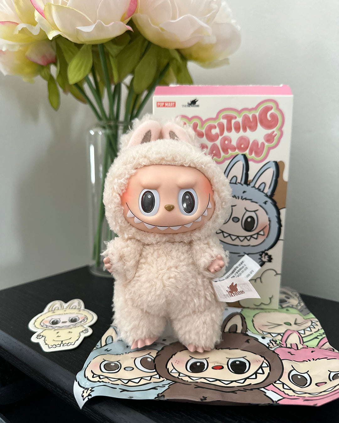 Plush toy with a character design on a table with a decorative box and flowers in the background.