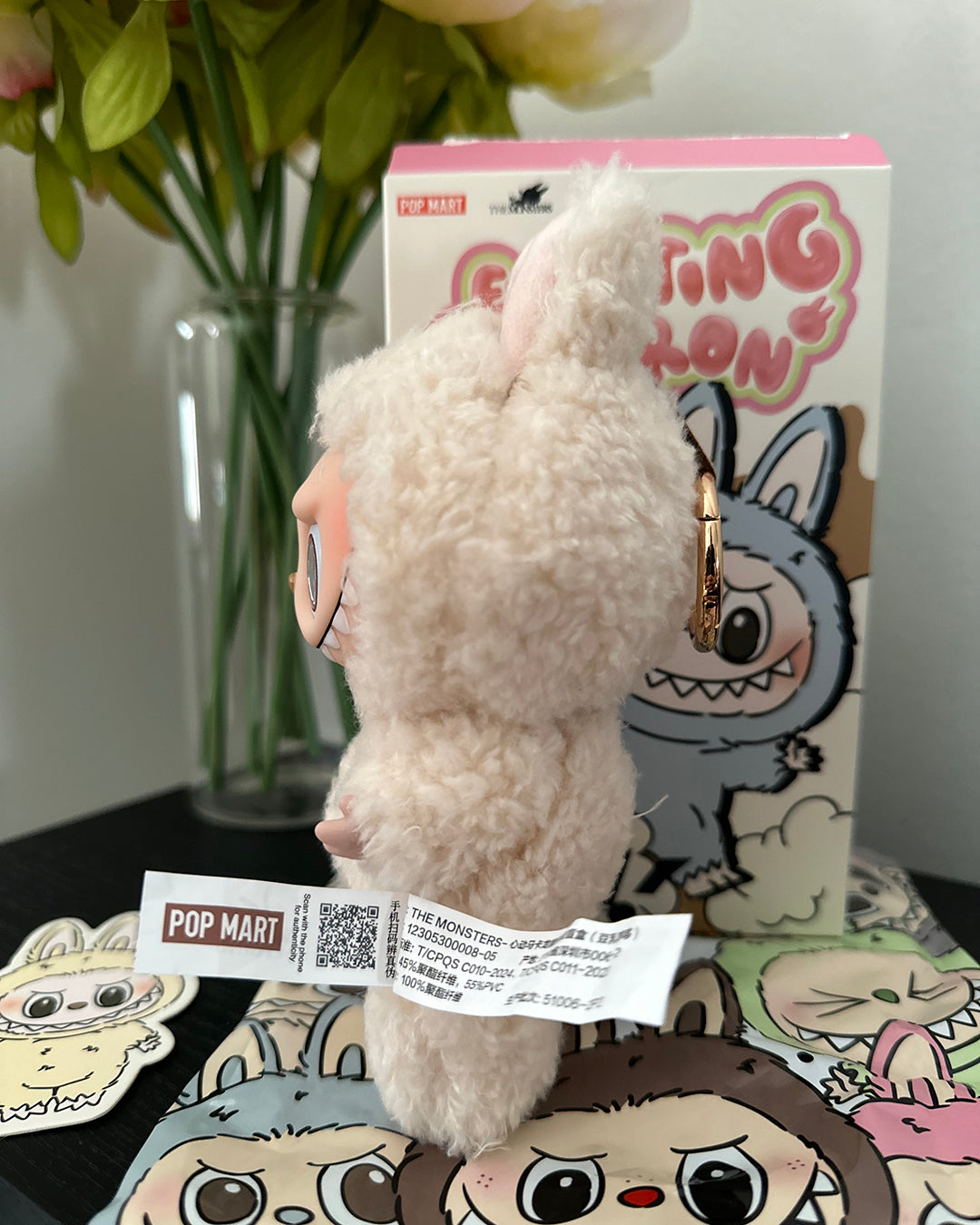 Plush toy with a keychain attached to a Pop Mart box, with flowers in the background.