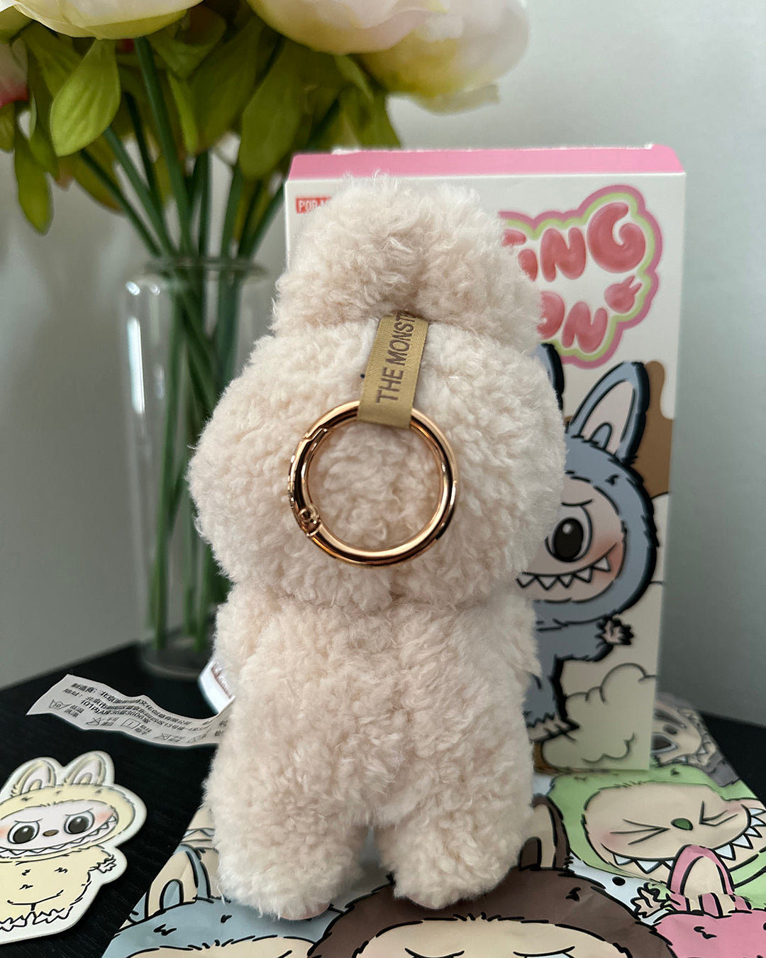 White fluffy keychain with gold ring on a table with cartoon character cards and flowers in the background