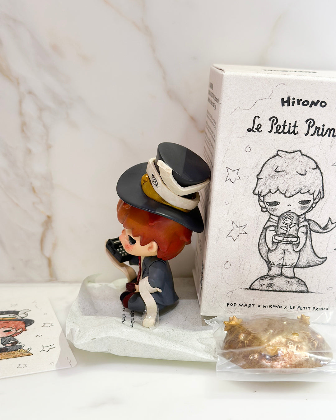 Small figurine with a hat next to a box labeled 'Hirono Le Petit Prince' on a white surface.