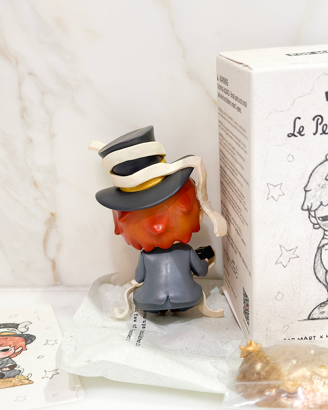 Small figurine with a hat next to a box labeled 'Le Petit Prince' on a white surface.