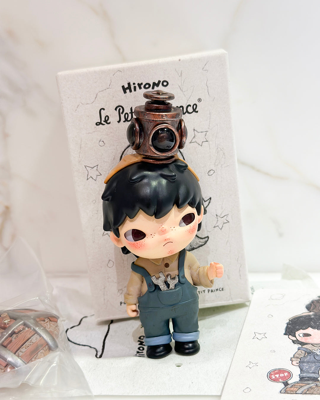 Small figurine with a lantern on its head, standing in front of a branded box.