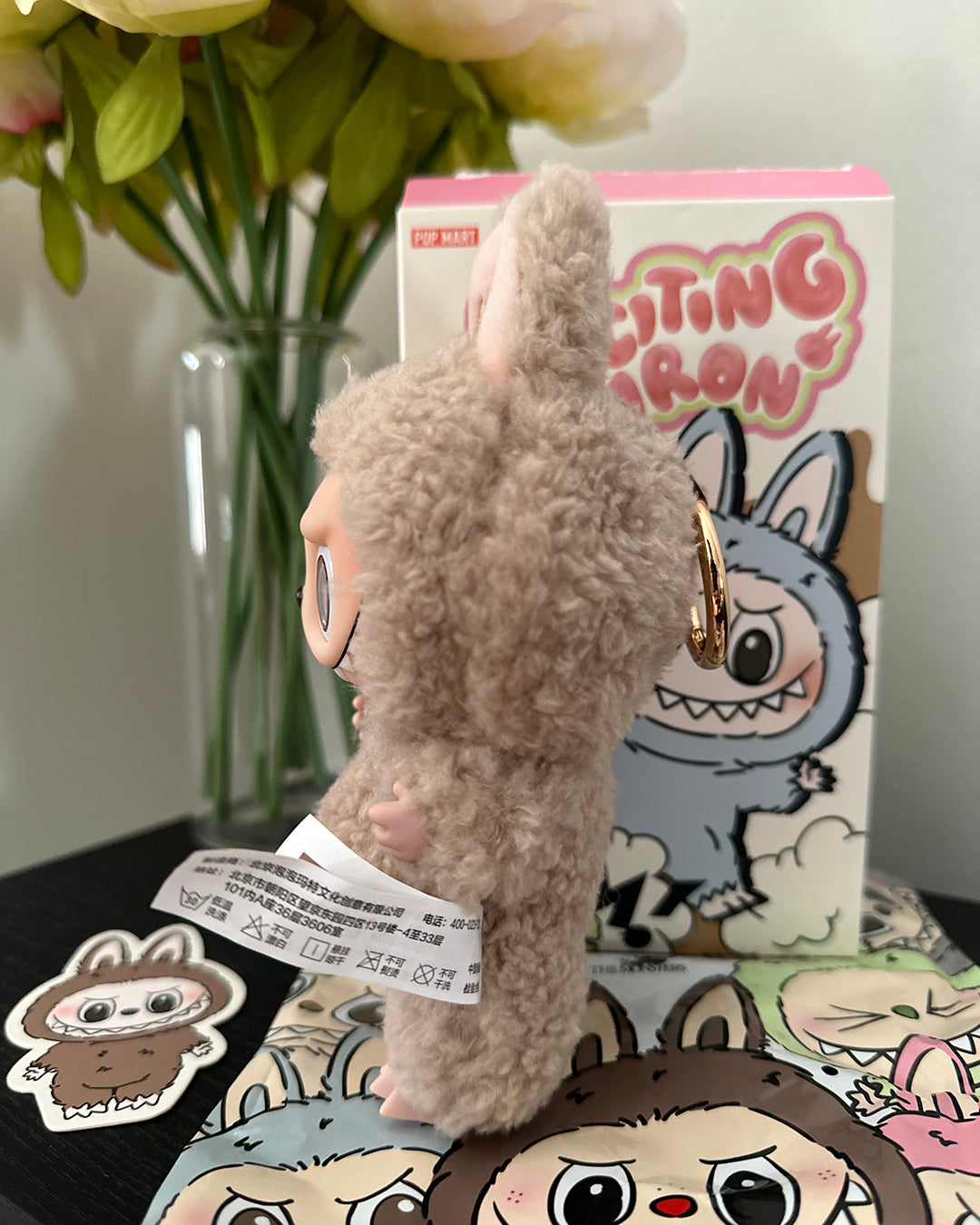 Plush toy with a keychain attached, sitting on a surface with a box and flowers in the background.