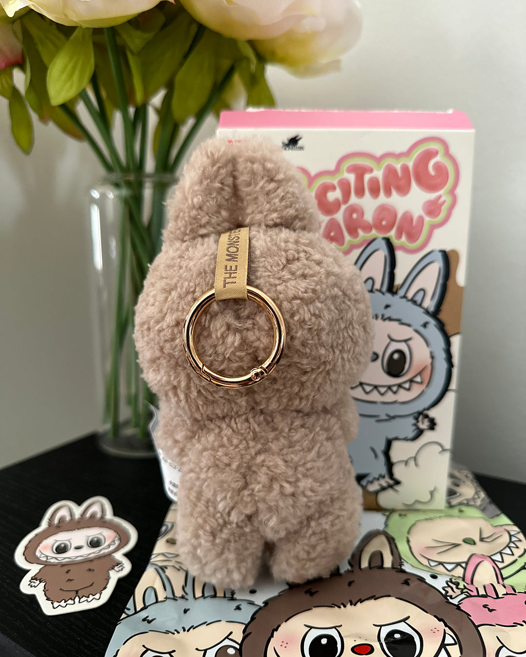 Fluffy keychain with a gold ring on a surface with a box and flowers in the background