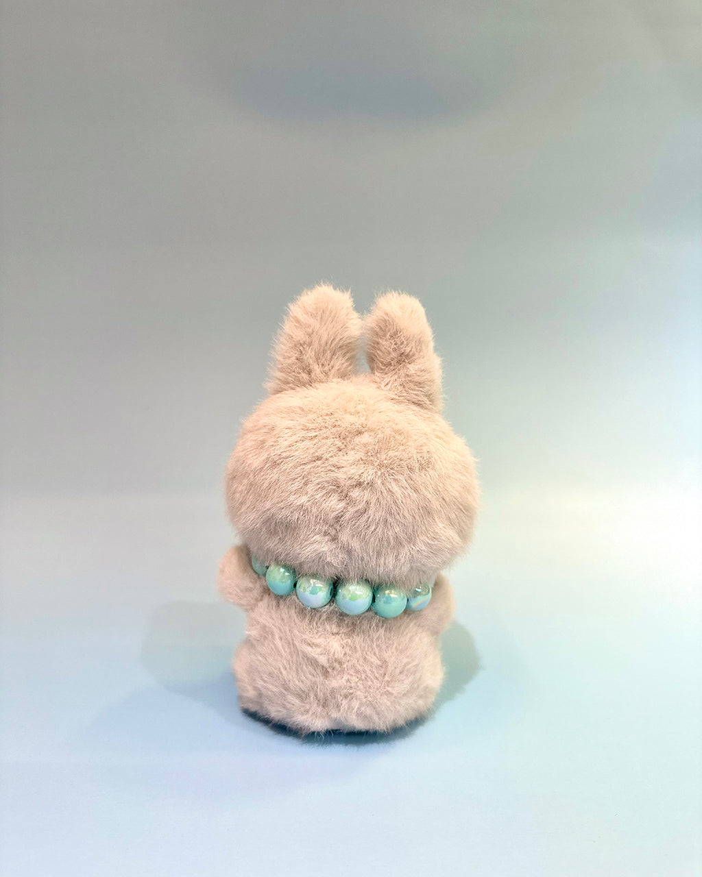 Fluffy beige plush toy with a green beaded necklace on a light gray background