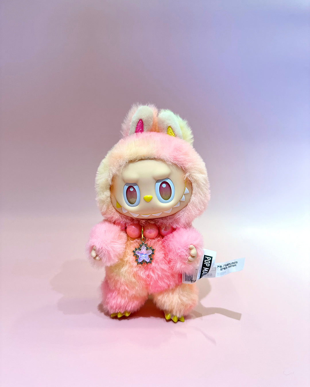 Plush toy with pink and white fur, colorful eyes, and a small tag on a light gray background