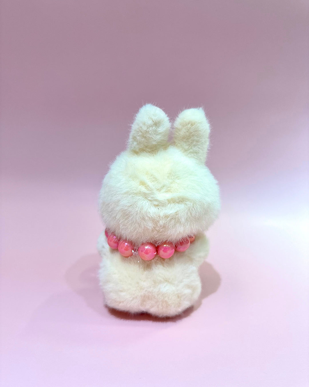 White fluffy toy with a pink beaded collar on a pink background