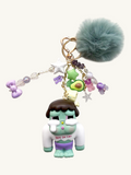 Keychain with character figure, avocado charm, and fur ball on a white background