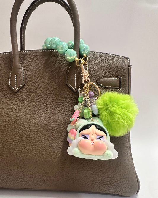 Decorative keychain with a cartoon character and green pom-pom attached to a brown handbag.