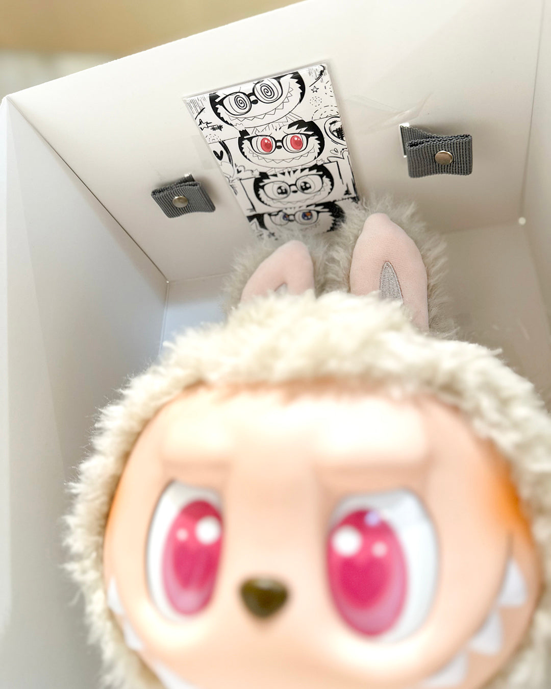 Close-up of a plush toy with large eyes and a small box on top, set against a neutral background.