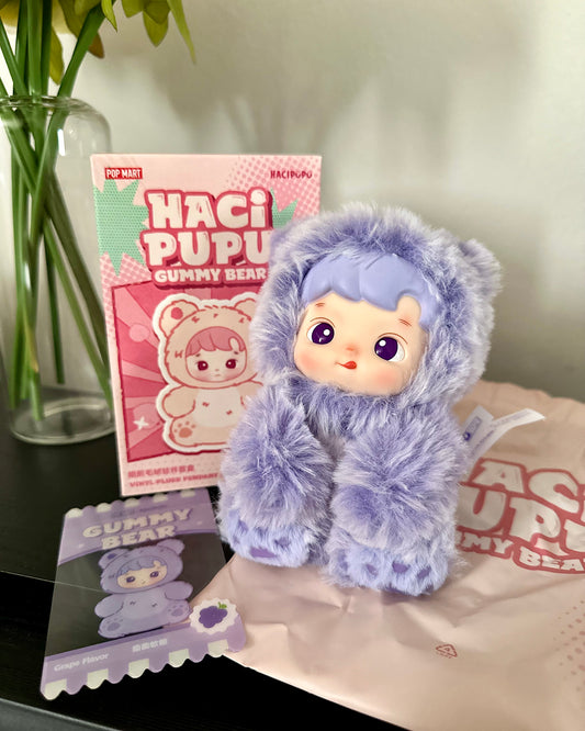 Plush toy with purple fur and a card featuring 'Haci Pupu' on a surface.