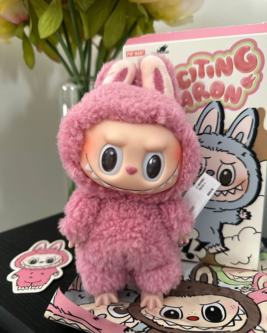 Pink plush toy with bunny ears on a surface with branded packaging in the background.