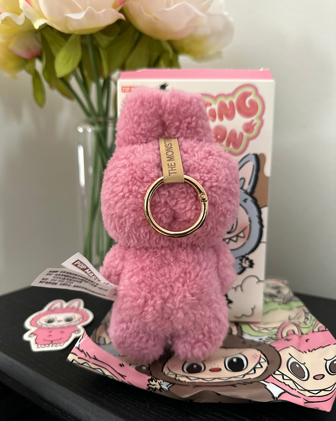 Pink fluffy keychain with a gold ring on a decorative surface with flowers and packaging in the background.