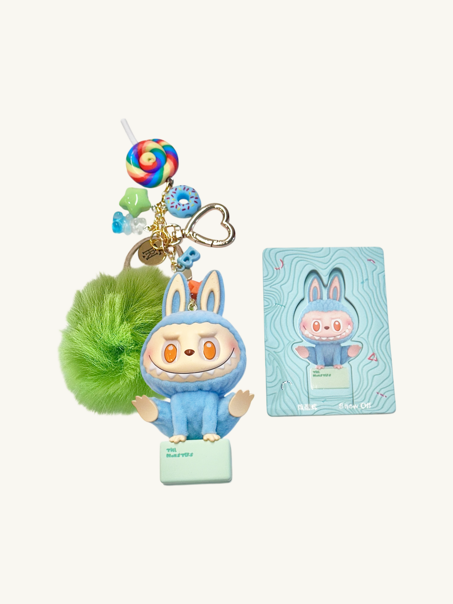 Blue cartoon character keychain with a green pom-pom and a lollipop charm, next to its packaging on a white background.