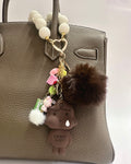 Decorative keychain with a fluffy pom-pom and small figure attached to a brown leather handbag.