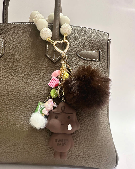 Decorative keychain with a fluffy pom-pom and small figure attached to a brown leather handbag.