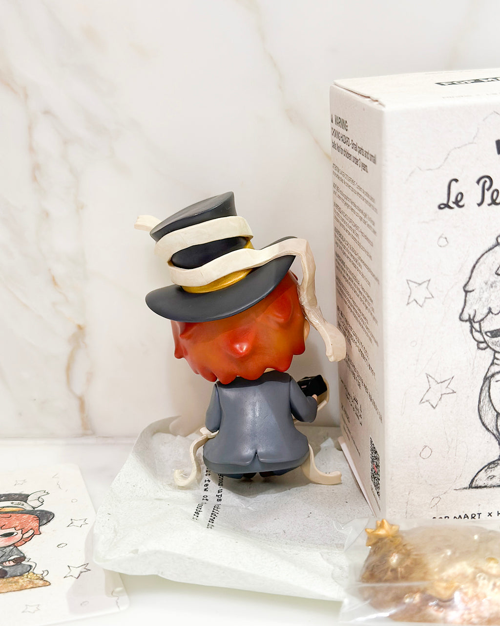 Small figurine with a hat next to a box labeled 'Le Petit Prince' on a white surface.