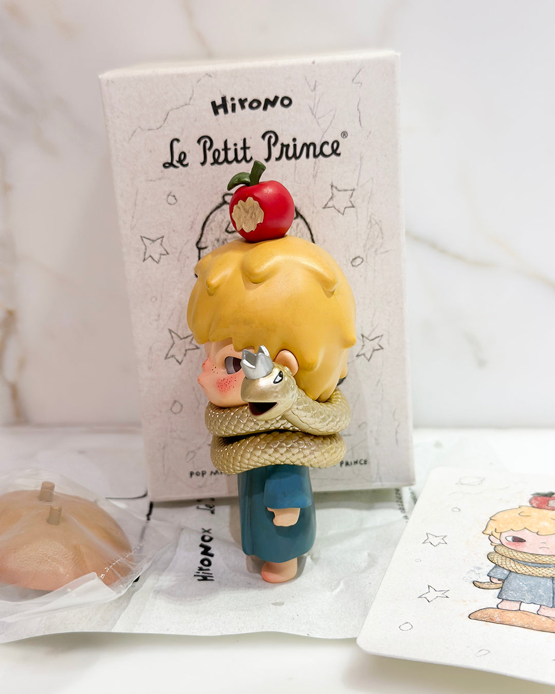Figurine of Le Petit Prince with an apple on his head, standing in front of a branded box.