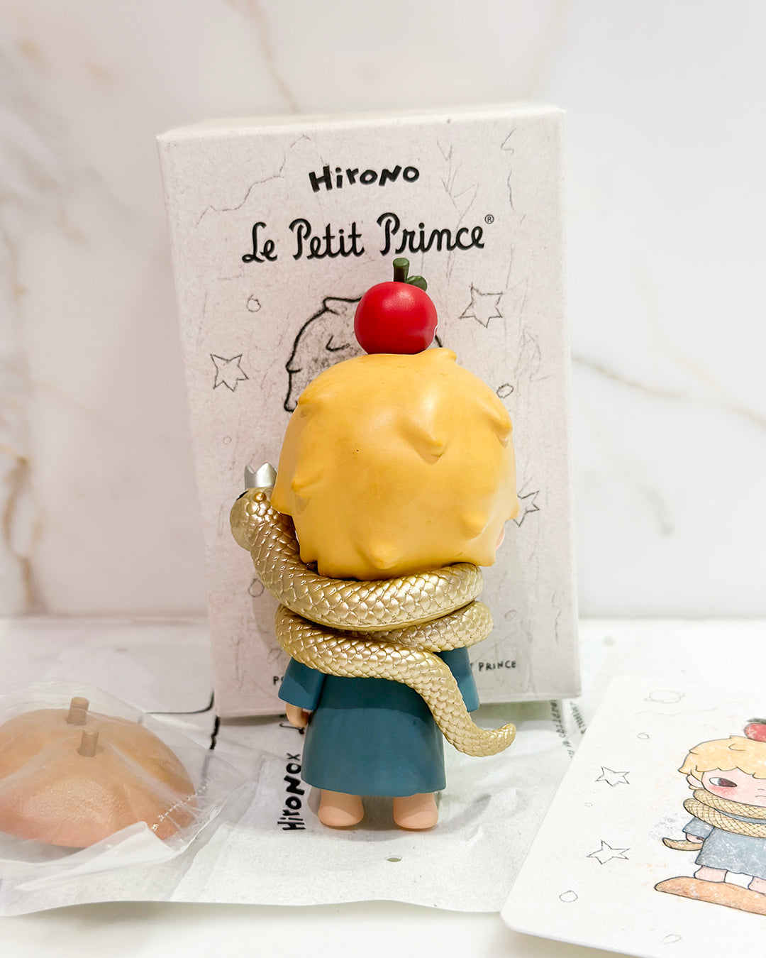 Toy figure of a character from 'Le Petit Prince' with a snake around its neck, standing in front of a branded box.